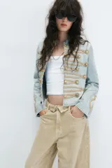 Chaqueta de mezclilla clara estilo militar, con cuello redondo, manga larga y detalles de cordones y botones dorados en el frente.