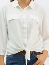 Camisa blanca de rayón con textura sutil, cuello con solapa, cierre frontal con botones y bolsillos simulados en el pecho. Presenta mangas 3/4 regulables con presilla y un diseño con nudo en el ruedo frontal.