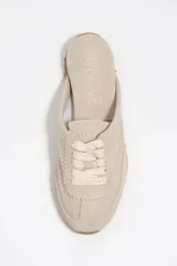 Zapatilla estilo mula de color beige, confeccionada en una combinación de algodón y serraje vacuno, con suela de caucho con tachuelas texturizadas y costuras decorativas visibles en la parte superior.