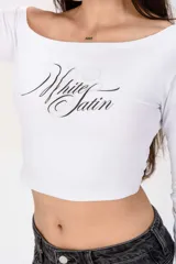 Remera crop blanca de manga larga, con escote asimétrico estilo off-shoulder y estampa frontal con la frase "White Satin" en letra cursiva negra.