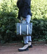 Bolso tipo shopper color verde oliva, con doble correa de mano en cuero negro y detalles de cintas verticales con logo estampado. Cuenta con cierre superior, bolsillos laterales y un diseño espacioso ideal para uso diario.