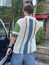 Camisa de manga corta tejida a crochet, con cuello polo y botones frontales. Presenta un diseño de rayas verticales en blanco, azul, verde y marrón.