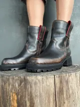 Bota de cuero con efecto desgastado en tonos marrones y negros, con suela track robusta y cierre lateral. Presenta detalles en color rojo en la parte superior de la caña.