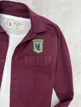 Sobrecamisa bordeaux de gabardina con cuello clásico, cierre frontal abotonado, dos bolsillos con solapa abotonada en el pecho y apliques bordados en el pecho y el hombro. Corte recto y mangas largas con puños abotonados.