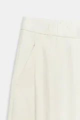 Pantalón fluido de color blanco, de tiro alto con trabillas y pliegues en el delantero. Cuenta con bolsillos laterales, cierre frontal con cremallera, botón interior y gancho metálico.