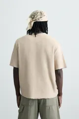 Camiseta de punto color beige claro, corte boxy fit, con cuello redondo y manga corta. Presenta un estampado central en tono similar que simula un efecto spray y acabados irregulares en los bordes.