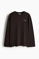 Remera de manga larga oversize confeccionada en punto de algodón suave, color marrón oscuro, con cuello redondo ribeteado, hombros caídos y pequeño estampado tipográfico en el pecho.