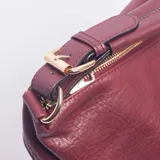 Cartera tipo bolso de color bordó, con textura granulada y detalles de dos correas decorativas con hebillas metálicas doradas en el frente. Cuenta con cierre superior, asa de hombro fija y correa larga ajustable y extraíble.