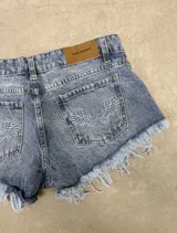 Short de denim azul con efecto deslavado, tiro medio y dobladillo desmechado con flecos. Presenta bolsillos traseros con bordado en forma de alas y etiqueta de cuerina de la marca Ask Denim en la pretina.