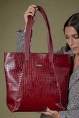 Bolso grande tipo tote de cuero con textura efecto croco en color bordó. Presenta detalles de costuras trenzadas verticales en el frente, doble asa de hombro y placa metálica decorativa con logo. El interior está forrado y cuenta con compartimentos organizadores.