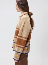 Pullover de punto con textura aterciopelada, diseño oversize, cuello polo con cierre frontal parcial y detalles de pespuntes en contraste. Presenta rayas horizontales en tonos tierra y un parche bordado con la letra M en el pecho.