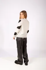 Sweater de punto color blanco con estampado de manchas negras irregulares y detalles de tachas metálicas sobre las mismas. Presenta cuello redondo y puños acanalados.