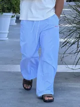 Pantalón de corte ancho y holgado, con diseño de rayas verticales finas en color azul y blanco. Presenta cintura elástica con cordón ajustable y un pequeño bordado en color rojo en la parte superior.