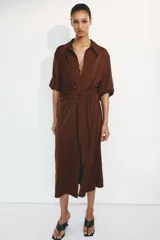 Vestido midi camisero de sarga liviana con brillo satinado, color marrón, de manga larga, con hombros caídos, botonadura frontal y puños sencillos. Presenta paneles drapeados cruzados en la cintura con tiras para atar al costado.
