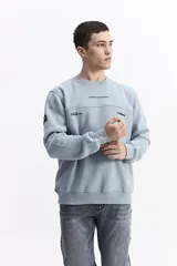 Buzo de algodón color celeste, con cuello redondo, puños y bolsillos laterales. Presenta estampados con texto en el pecho y un parche en la manga.