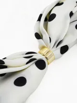 Pañuelo cuadrado de tela ligera color negro con estampado de lunares blancos y borde con franja blanca. Incluye un anillo metálico dorado para sujetar y estilizar el pañuelo.