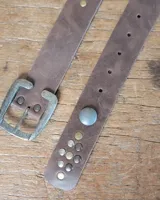Cinto de cuero marrón con hebilla metálica de estilo envejecido y detalle de tachas metálicas en diferentes tonos en el extremo.
