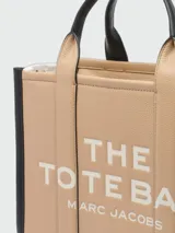 Cartera tipo tote bag Marc Jacobs modelo The Medium Leather Tote Bag, color marrón tostado con detalles en negro, confeccionada en cuero con las inscripciones "THE TOTE BAG" y "MARC JACOBS" estampadas en el frente. Cuenta con asas superiores y correa ajustable y desmontable para el hombro.