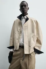 Cazadora regular fit de algodón color beige, con cuello subido y forro interior a cuadros. Presenta cierre frontal de cremallera oculta por solapa con botones a presión, bolsillos de vivo en la cadera y puños con botón.