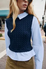 Camisa de popelín celeste con cuello solapa y manga larga, combinada con un chaleco de punto trenzado en color azul marino superpuesto. Presenta un diseño de capas con cierre en la espalda mediante botones ocultos.