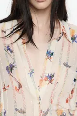 Camisa oversize de tejido fantasía color crema con estampado de pájaros y flores en tonos azules, amarillos, naranjas y rojos. Presenta finas rayas verticales en colores pastel y lentejuelas distribuidas por toda la prenda. Tiene cuello camisero, cierre frontal con botones y mangas cortas rectas con un ligero vuelo. Incluye un bolsillo de plastrón en el pecho izquierdo.