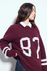 Sweater de tejido de punto color blanco con estampado numérico '87' en negro en el frente y detalles de rayas horizontales en las mangas. Presenta cuello redondo y un calce relajado.
