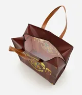 Bolsa de compras marrón con diseño de maleta y estampa frontal del escudo de Hogwarts. Cuenta con asas dobles de mano y cierre superior.