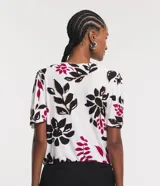 Blusa de viscosa con escote en V y mangas cortas abullonadas. Presenta un estampado de follaje y flores en tonos negro, fucsia y beige sobre fondo blanco.