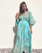 Túnica larga de estilo boho con escote en V profundo, mangas amplias tipo kimono y detalle de cordones con borlas en el frente. Presenta un estampado de arabescos y detalles en hilo metalizado.