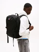 Mochila estilo retro de la marca Tommy Jeans, color negro, inspirada en modelos utilitarios. Cuenta con compartimento principal con cierre de cremallera, bolsillo exterior con cremallera, panel delantero con textura acolchada y parche con el logo de la marca en la parte superior. Incorpora correas ajustables, asa superior y detalles de tiras trenzadas.