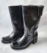 Bota de cuero negro con diseño 2 en 1, que incluye una polaina de gamuza desmontable. Presenta una base de bota de cuero liso y una caña alta de gamuza, ofreciendo versatilidad para diferentes estilos.