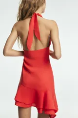 Vestido corto de tejido fluido en color rojo, con diseño de cuello halter y detalle de frunces laterales. Presenta un bajo asimétrico acabado en volantes y cierre lateral con cremallera oculta.