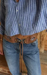 Cinturón de cuero vacuno color marrón, con diseño artesanal cosido a mano, cierre frontal con argollas metálicas y tiras de ajuste.