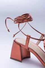 Sandalias de cuero color cobre con taco cuadrado y tira fina sobre los dedos. Presentan una flor grande en el empeine y tiras finas para atar al tobillo.