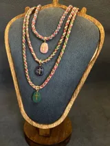 Collar de diseño artesanal compuesto por cordones trenzados multicolor en tonos verdes, rosados y rojizos. Presenta un dije de piedra ovalada con una cruz dorada aplicada en el centro.