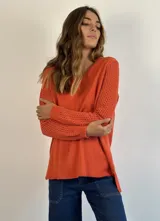 Buzo de punto color naranja con cuello en V y mangas largas con textura calada. Presenta un diseño holgado con tajos laterales en el ruedo.