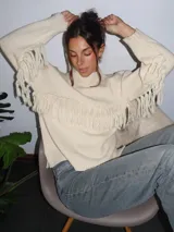Sweater tejido a mano en lana merino, color crudo, con cuello a la base, mangas holgadas y detalle de flecos aplicados en el frente y las mangas.