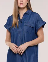 Vestido camisero corto de jean color azul, con cuello camisero, escote en V y botones frontales parciales. Tiene mangas cortas con dobladillo y bolsillos con solapa en el pecho.