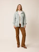 Campera de corte recto y estilo funcional, color beige, con capucha integrada, cierre frontal con botones a presión y bolsillos delanteros amplios. Presenta un diseño minimalista con cordones ajustables en la capucha.