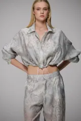 Conjunto de dos piezas compuesto por una chaqueta corta tipo crop con cordón ajustable en el ruedo y un pantalón de corte recto. Ambas prendas están confeccionadas en tela de organza con estampado floral tipo damasco en tonos grisáceos.