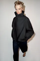 Cazadora corta de color negro con cuello subido cruzado, manga larga con puños anchos ajustados con trabilla y botón, y cierre frontal cruzado con botones ocultos.