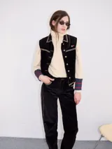 Chaleco de pana negro con cierre frontal de botones metálicos dorados. Presenta detalles de costuras en contraste en el cuello, cartera y canesú, e incluye un pequeño parche con forma de bandera en el pecho.