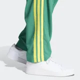 Pantalón deportivo Adidas de corte estándar, color verde con las icónicas tres rayas laterales en color amarillo. Presenta logo de la marca bordado en violeta y bolsillos laterales.