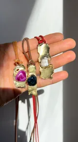 Collar artesanal con dije de base metálica irregular y piedra central roja. Presenta una estructura de tiento de gamuza marrón con detalles de tubos metálicos dorados.