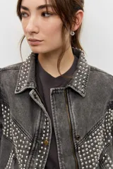 Campera de jean color gris con efecto acid wash, corte clásico y cierre frontal de botones metálicos. Presenta cuello camisero, bolsillos ribeteados y está decorada con apliques de tachas plateadas en el cuello, el canesú frontal, los bordes de la botonera, los bolsillos y el ruedo.