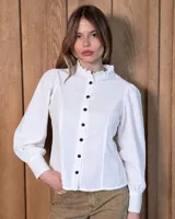 Camisa de jean blanca con cuello alto con volados, cierre frontal con botones oscuros y mangas largas abullonadas con puños ajustados.