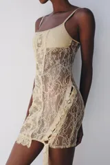 Vestido corto semitransparente tipo corsetero color beige, confeccionado con encaje y detalles de varillas interiores que estilizan la figura. Presenta escote redondo, tirantes finos ajustables y bajo asimétrico con una lazada lateral de satén.