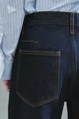 Jeans de tiro bajo y pernera ancha, confeccionados en denim azul oscuro con pespuntes en contraste. Cuentan con un diseño de cinco bolsillos, cierre frontal con cremallera y botón metálico, y destacan por su amplio doblando en el bajo que deja ver el revés de la tela con detalles de costura selvedge.