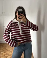 Sweater de tejido bremer con diseño de rayas horizontales en tonos vino, gris y blanco. Presenta cuello polo con solapa abotonada, mangas largas y corte holgado.