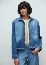 Campera corta de denim azul con efecto desgastado. Tiene cuello clásico, cierre frontal con botones, dos bolsillos delanteros y terminaciones desflecadas.